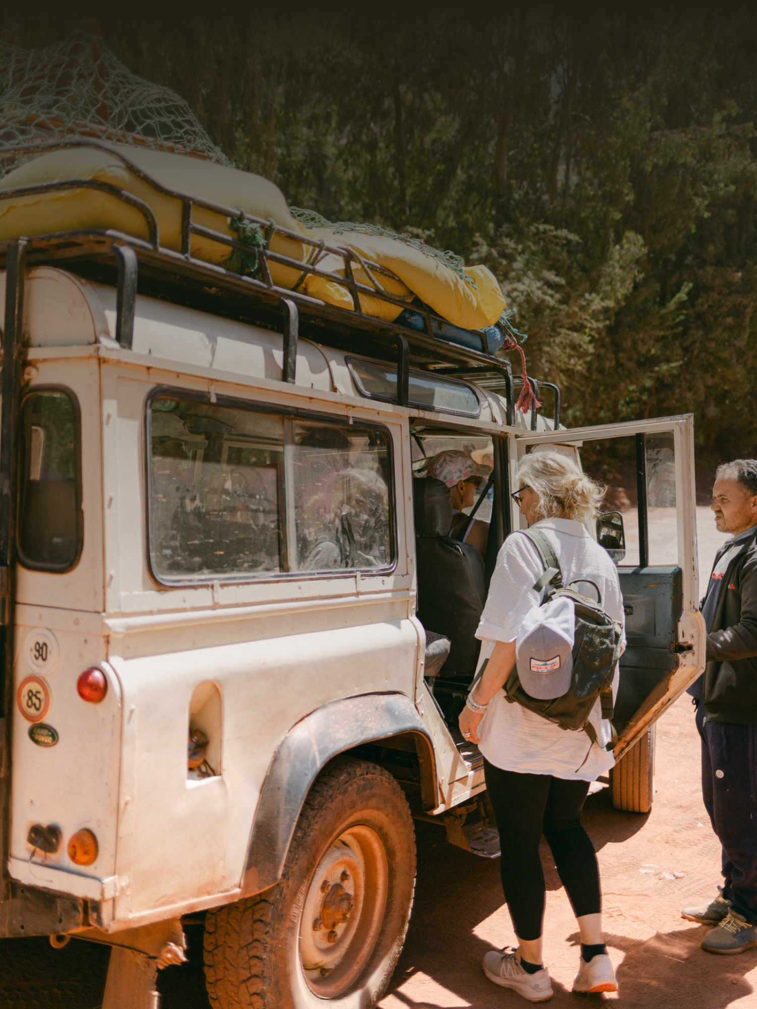 a woman getting into a white jeep with luggage on top. the surroundings suggest a nature setting.